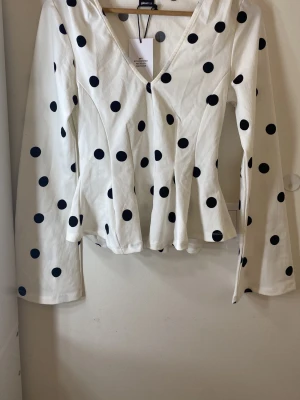 Vit prickig blus från Gina Tricot - Trendig vit blus med svarta stora prickar från Gina Tricot. Blusen har v-ringning, figurnära passform och långa ärmar. Tillverkad i ett mjukt och lätt tyg som ger en snygg siluett. Perfekt för dig som gillar retrovibbar och lekfull stil.