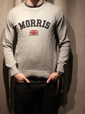 Grå stickad tröja från Morris - Snygg grå stickad tröja från Morris med svart broderad logotyp och brittisk flagga på bröstet. Tröjan har rund halsringning, ribbstickade muddar och är perfekt för en stilren look. Står S men passar även M utan problem.Tröjan har inga defekter. Hör av dig vid frågor🌟