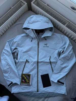 Ljusblå Arc'teryx vindjacka Gore-Tex - Snygg ljusblå vindjacka från Arc'teryx med huva och vattentätt Gore-Tex-material. Jackan har två snedställda dragkedjefickor, justerbar huva och diskret logga på bröstet. Perfekt för blåsiga dagar och outdoor-äventyr.