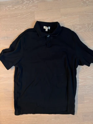 Svart pikétröja från COS - Klassisk svart pikétröja från COS med korta ärmar och krage med två knappar. Tillverkad i mjuk bomull som känns skön mot huden. Perfekt till jeans eller shorts för en clean och enkel stil.