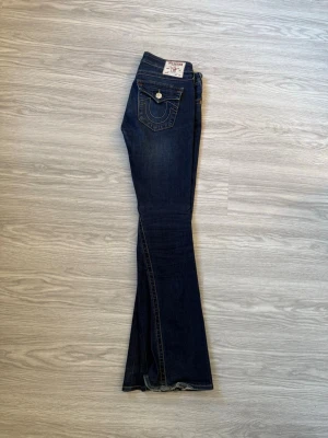 True Religion bootcut jeans mörkblå - Mörkblå bootcut jeans från True Religion med klassiska kontrastsömmar och snygga detaljer på bakfickorna. Modellen heter Joey och har låg midja samt fem fickor. Perfekta för dig som gillar en avslappnad men trendig look.