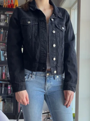 Svart jeansjacka med silverknappar - Snygg svart jeansjacka med klassisk krage och silverfärgade knappar framtill. Två bröstfickor med lock och knapp, samt justerbara knappar i midjan bak. Kortare modell som passar perfekt till jeans eller kjol.