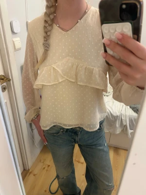Prickig cremefärgad blus med volang - Söt cremefärgad blus med små prickar och volangdetalj framtill. Blusen har v-ringning och är gjord i ett tunt, transparent tyg som ger en luftig känsla. Långa ärmar med lätt puff och resår vid ärmslut. 