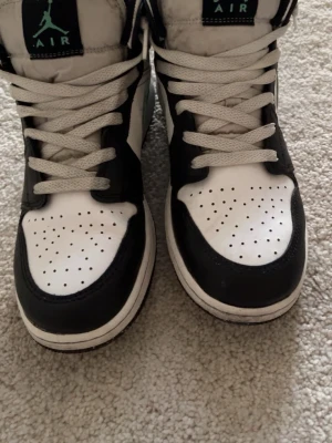 Air Jordan 1 Mid svart/vit/turkos - Säljer ett par Air Jordan 1 Mid sneakers från Nike i svart och vitt med turkosa detaljer på swoosh och logga. Skorna har klassisk hög siluett, perforerad tåbox och vita skosnören. Tillverkade i läder och textil för en clean och sportig look.