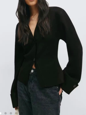 Svart kofta med vida ärmar - Stilren svart kofta med v-ringning och två knappar framtill. Koftan har vida, långa ärmar med breda manschetter och knappdetalj. Perfekt till jeans eller kjol för en clean look.