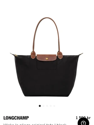 Longchamp svart Le Pliage handväska - Klassisk svart handväska från Longchamp Le Pliage med bruna läderhandtag och lock. Väskan har en enkel, rymlig design i nylon med guldfärgad knappdetalj och dragkedja. Perfekt för dig som gillar stilren och tidlös look. Storleken är 31x30x19cm💗( knappast använd och ser helt ny ut)