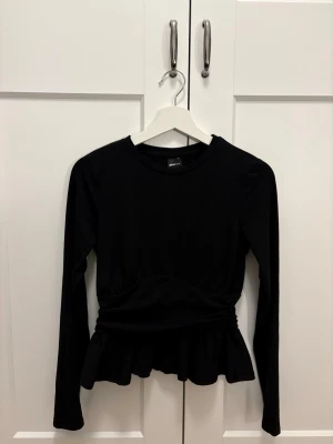 Svart volangtopp från Ginatricot - Supersnygg svart topp från Ginatricot med långa ärmar och volang nertill. Modellen har en markerad midja och rund halsringning. Perfekt att styla med jeans eller kjol. Använd endast en gång