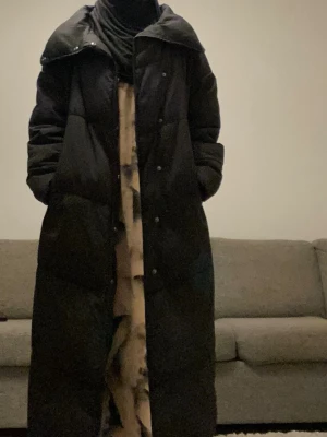 Lång svart dunjacka med hög krage - Säljer en lång svart dunjacka med hög krage och breda kanaler. Jackan har en oversized passform, tryckknappar framtill och stora fickor på sidorna. Perfekt för kalla vinterdagar och riktigt mysig att bära.