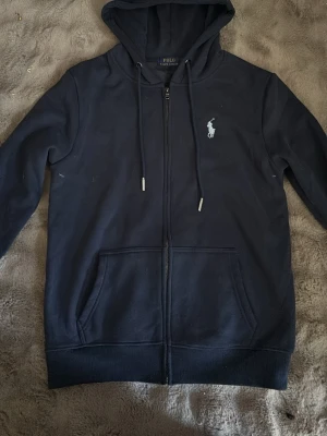 Mörkblå hoodie från Polo Ralph Lauren - Snygg mörkblå hoodie med dragkedja från Polo Ralph Lauren. Klassisk design med vit broderad logga på bröstet, huva med dragsko och stora fickor framtill. Perfekt för en chill och stilren look.