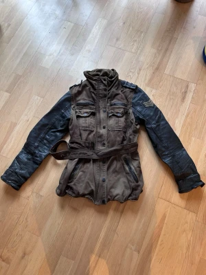 Brun och svart skinnjacka med bälte - Cool brun och svart skinnjacka med bälte i midjan och quiltade detaljer på ärmarna. Jackan har fyra fickor framtill, hög krage och axelklaffar för en tuff look. Perfekt för dig som vill sticka ut med en unik jacka. Strl 40 men jag använder 38 i vanliga fall så liten i storlek.