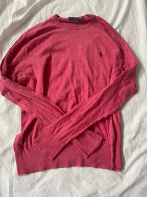 Rosa stickad tröja från Polo Ralph Lauren - Säljer en snygg rosa stickad tröja från Polo Ralph Lauren med klassisk rund halsringning och den ikoniska svarta loggan broderad på bröstet. Tröjan har långa ärmar och ribbade muddar vid ärmslut och nederkant. Perfekt för lager på lager under kyliga dagar.