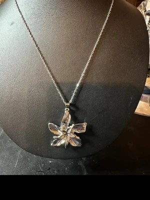 Silverfärgat halsband med blomma - Snyggt halsband i silverfärg med en stor, detaljerad blomma som hänge. Kedjan är tunn och länkarna är små, vilket ger ett elegant intryck. Hänget har en unik, handgjord känsla med tydliga ådror i blombladen. Perfekt för dig som gillar naturinspirerade smycken.