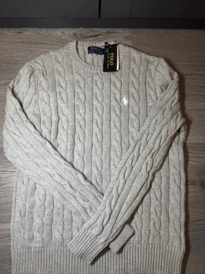 Grå kabelstickad tröja Polo Ralph Lauren - Snygg grå kabelstickad tröja från Polo Ralph Lauren med rund halsringning och klassisk vit broderad logga på bröstet. Tröjan har ribbade muddar vid ärmslut och nederkant och är tillverkad i mjukt stickat material.