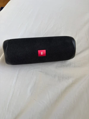 Svart JBL bärbar högtalare - Snygg svart bärbar högtalare från JBL med cylinderformad design och robust tygmaterial. Högtalaren har tydliga knappar på ovansidan för enkel styrning och en röd JBL-logga på framsidan. Perfekt för att ta med sig musiken överallt.