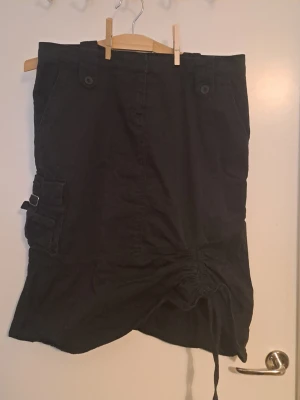 Svart cargo kjol  - Svart kjol med cargofickor på sidorna. Tråd fram sim man kan dra åt för scrunch look. Väldigt fin. Är mer svart än på bilden. Str 40🩷 Hittar inga defekter