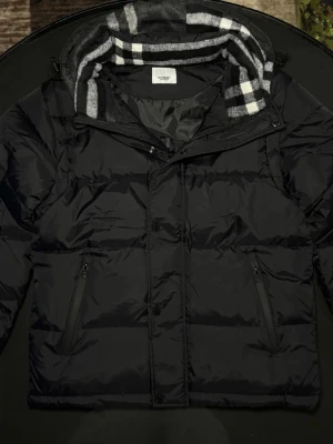 Svart burberry jacka - Stilren svart dunväst från Burberry  med avtagbara ärmar och huva. Västen har två dragkedjefickor och en fodrad huva med rutigt mönster i grått, vitt och svart. Perfekt för lager-på-lager och streetstyle.