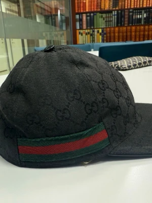 Svart keps från Gucci med GG-mönster - Snygg svart keps från Gucci med klassiskt GG-monogram över hela kepsen. På sidan finns en ikonisk grön och röd rand. Kepsen har böjd skärm och är tillverkad i ett tygmaterial som känns lyxigt och stilrent. Perfekt för dig som vill ha en exklusiv accessoar. Pris går o diskutera ( den är äkta )