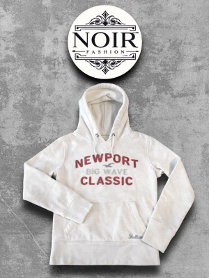 Vit hoodie från Hollister - Vit hoodie med stor huva och snörning från Hollister. Tryck i rött och grått med texten 'Newport Big Wave Classic' på bröstet. Mjuk bomullsmix och klassisk känguruficka framtill. Perfekt för en avslappnad och cool look.