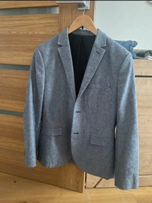 Grå kavaj från HM med mockadetalj - Snygg grå kavaj med skön slim fit och snygga bruna mockalappar på armbågarna. Kavajen har två framfickor, en bröstficka och fyra knappar vid ärmsluten. Perfekt för en schysst look.