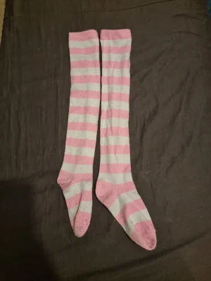 Rosa och vita randiga knästrumpor - Säljer ett par långa knästrumpor med breda rosa och vita ränder. Strumporna är mjuka och stretchiga, perfekta för att ge en söt touch till din outfit. Inget synligt märke på strumporna. Passar dig som gillar färgglada detaljer.
