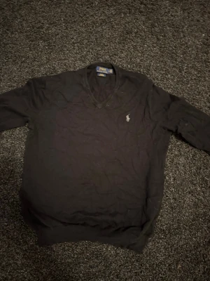 V ring tröja från Polo Ralph Lauren - Klassisk svart tröja från Polo Ralph Lauren med V-ringning och den ikoniska loggan broderad på bröstet. Tröjan har långa ärmar och ribbade muddar vid ärmslut och nederkant. Perfekt för en stilren och enkel look.