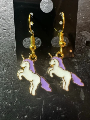 Lila enhörningsörhängen - Säljer ett par gulliga örhängen med enhörningar i vitt och lila emalj. Hänget har guldfärgade detaljer och krokar. Perfekt för dig som älskar magiska accessoarer och vill sticka ut lite extra.