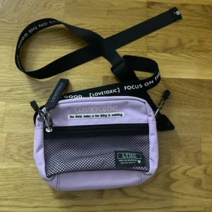 Ljuslila handväska från Lovetoxic - Snygg ljuslila handväska från Lovetoxic med svart justerbar axelrem och coola textdetaljer. Väskan har ett nätfack framtill med dragkedja och en genomskinlig ficka på baksidan. Perfekt för dig som gillar streetstyle och vill sticka ut.