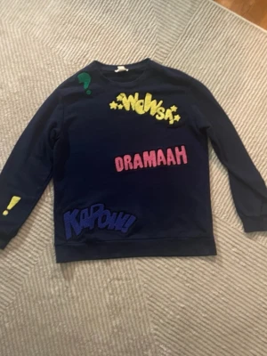 Mörkblå sweatshirt med färgglada tryck - Cool mörkblå sweatshirt med broderade och tryckta detaljer som WOWSA!, DRAMAAH, KAPOW!, utropstecken, stjärnor och hjärta. Tröjan har rund hals och långa ärmar. Perfekt för dig som gillar statement-plagg och serietidningsstil.