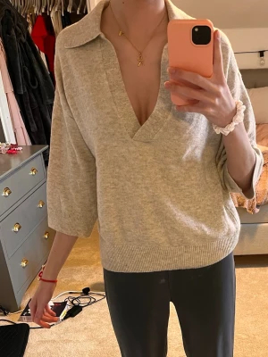 Beige stickad tröja med krage - Snygg beige stickad tröja med djup v-ringning och krage. Tröjan har halvlånga ärmar och ribbstickade muddar vid ärmslut och nederkant. Perfekt för en avslappnad och trendig look.