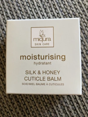 Miqura Silk & Honey Cuticle Balm - Nagelbandsbalsam från Miqura med silke och honung som återfuktar och mjukgör nagelbanden. Innehåller sheasmör, mandelolja och jojobaolja. Kommer i en vit förpackning med guldtext. Perfekt för dig som vill ta hand om dina nagelband.