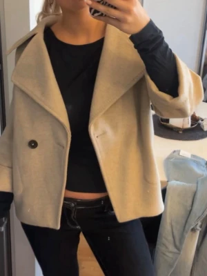 Beige ullkappa med vida ärmar - Stilren beige kappa i ullblandning med oversized passform och vida trekvartsärmar. Kappan har bred krage, en knapp framtill och två fickor. Perfekt för lager-på-lager och en trendig look.