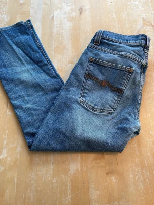 Blå jeans från Nudie Jeans - Säljer ett par klassiska blå jeans från Nudie Jeans med snygga slitningar och kontrastsömmar på bakfickan. Modellen Grim Tim. Jeansen har fem fickor och är gjorda i bomull med en cool vintage-look. Perfekta för dig som gillar en avslappnad stil. Storlek 31w 32l men sitter lite smått. Några mini fläckar på framsidan. 