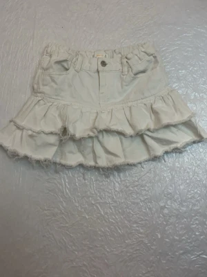 Vit volangkjol med råa kanter - Supersöt vit kortkjol med dubbla volanger och råa kanter för en avslappnad look. Kjolen har fickor fram och bak, knapp och dragkedja i midjan samt hällor för bälte. Perfekt för dig som gillar en trendig och lekfull stil.