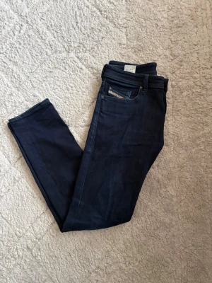 Mörkblå slim fit jeans från Diesel - Säljer ett par mörkblå slim fit jeans från Nudie Jeans. Klassisk femficksmodell med diskreta sömmar och knappgylf. Jeansen har en stilren look och är tillverkade i mjukt denimtyg med lite stretch för extra komfort. Se sista bild för storlek