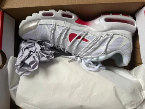 Nike Air Max Plus vita/röda sneakers - Nike Air Max Plus sneakers i vitt mesh med röda detaljer och synlig Air-sula. Skorna har en sportig siluett, vita snören och plastdetaljer längs sidorna. Perfekta för dig som gillar streetwear och vill sticka ut med klassisk Nike-design.