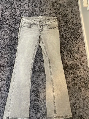 Grå bootcut jeans med tvättad look - Säljer ett par grå bootcut jeans med tvättad effekt och klassiska fem fickor. Jeansen har utsvängda ben och markerade sömmar för en cool vintagekänsla. Perfekta för dig som gillar retrostil och vill sticka ut lite extra.