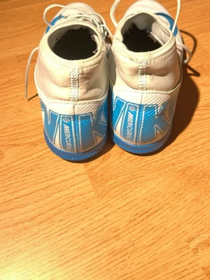 Nike Mercurial vita/blå fotbollsskor - Säljer ett par Nike Mercurial fotbollsskor i vitt och blått med snörning. Skorna har en låg profil och slitstark yttersula i blått gummi, perfekt för inomhusspel. Stora Nike-loggor på hälen och sidan. Tillverkade i syntetmaterial för bra passform och känsla.