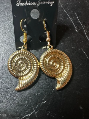 Guldiga spiralformade örhängen - Säljer ett par coola örhängen i guldtonad metall med spiralform som påminner om snäckskal. De har en unik, detaljerad struktur och hängs på krok. Perfekta för dig som vill sticka ut med accessoarer som har en retrovibe.