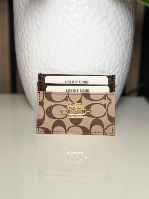 Coach brun korthållare i läder - Snygg korthållare från Coach i brunt läder med klassiskt monogrammönster och guldfärgad logotyp framtill. Perfekt för att organisera dina kort på ett stilrent sätt. Smidig och tunn design som enkelt får plats i fickan.