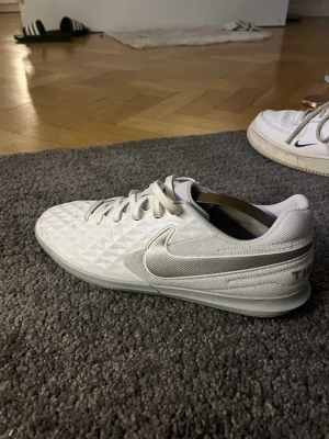 Nike Tiempo vita fotbollsskor - Snygga vita Nike Tiempo fotbollsskor med diskret silverfärgad swoosh och textur på ovandelen. Skorna har snörning och är tillverkade i syntetmaterial med meshpaneler för bra ventilation. Perfekta för dig som vill ha stilrena och bekväma skor på planen. OBS: saknar skorsnöret på höger sidan. 