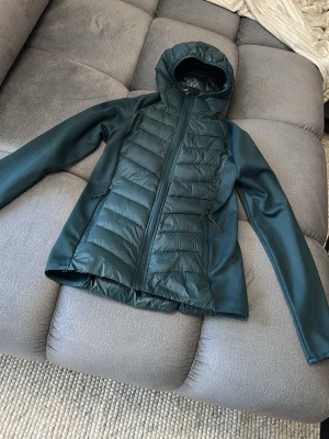 Mörkgrön pufferjacka med huva - Snygg mörkgrön pufferjacka med huva och dragkedja framtill. Jackan har quiltade partier på bröstet och ryggen samt släta ärmar. Två sidofickor med dragkedja. Perfekt för kalla vinterdagar och ger en sportig look.