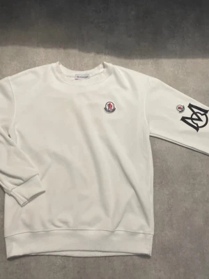 Mnclr tröja - Vit sweatshirt med rund halsringning och M-logga på bröstet. På ena ärmen finns en stor svart M-print och en liten logga. Tillverkad i mjukt bomullsmaterial och har ribbade muddar vid ärmslut och nederkant. Perfekt för en clean och stilren look.