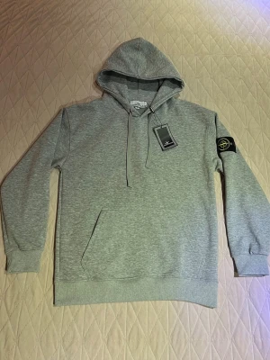 Grå hoodie från Stone Island - Snygg grå hoodie från Stone Island med klassisk logga på vänster ärm. Hoodien har dragsko i huvan, magficka och ribbade muddar. Tillverkad i mjuk bomullsblandning som är skön att ha på sig. Perfekt för en avslappnad streetwear-look. SITTER SOM EN S