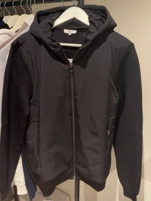 Svart vindjacka från ARNE med huva - Svart vindjacka från ARNE med huva och hel dragkedja framtill. Jackan har två fickor med dragkedja och är tillverkad i ett slätt syntetmaterial med stickade partier på ärmarna. Perfekt för blåsiga dagar och enkel att matcha.