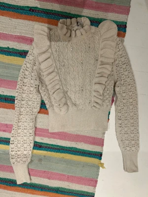 Beige stickad tröja med volanger och strass - Supersöt beige stickad tröja med höghalsad krage och dekorativa volanger framtill och baktill. Tröjan har ett fint hålmönster på ärmarna och strass på framsidan. Ribbstickad mudd vid midja och ärmslut.