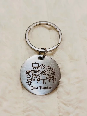 Nyckelring med gravyr 'Best Teacher' - Rund silverfärgad nyckelring i metall med graverad text 'Best Teacher' och en söt illustration av en lärare och flera barn. Perfekt accessoar till väskan eller nyckelknippan för att visa uppskattning till en favoritlärare.