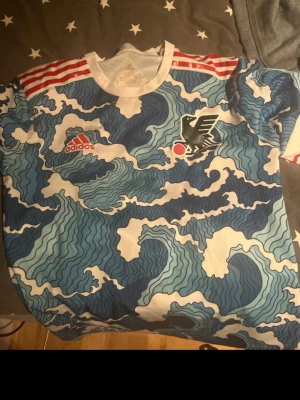 Adidas Japan tröja vågmönster M - Säljer en Adidas Japan tröja i storlek M med unikt blått och vitt vågmönster, röda ränder på axlarna och tryckt emblem på bröstet. Tillverkad i lätt och ventilerande material, perfekt för sport eller träning. Screenshot pga andra hemsidor…