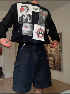 Svart croppad sweatshirt med patchar - Svart croppad sweatshirt med långa ärmar och flera stora patchar framtill i vitt, rött och grått med grafiska tryck och anarkisymbol. Tröjan har en edgy, alternativ vibe och är tillverkad i mjuk bomull. Perfekt för dig som gillar streetwear och statement-plagg.