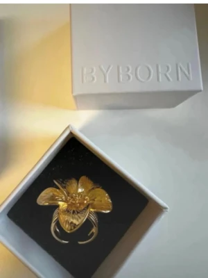 Guldfärgad blomring från Byborn - Large flower ring gold nypris 650kr Adjustable  18k guldpläterad mässing  Guldig ring Blomma Justerbar ring Guldring Handgjorda smycken Guldig ring från By Born. Unik ring från Byborn i guldfärgad metall med en stor, detaljerad blomma som motiv. Ringen har fem kronblad och en elegant, blank finish som verkligen fångar ögat. Perfekt för dig som vill sticka ut med ett statement-smycke.