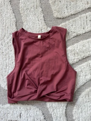 Ljusvinröd croppad träningstopp - Snygg ljusvinröd croppad träningstopp med twistad detalj framtill. Ärmlös modell med rund halsringning och söm mitt fram. Helt oanvänd. 